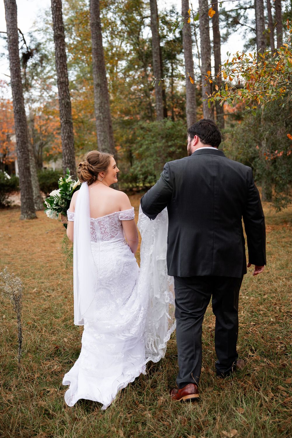 Wedding couple walking through a wooded area with the bride's dress trailing behind.