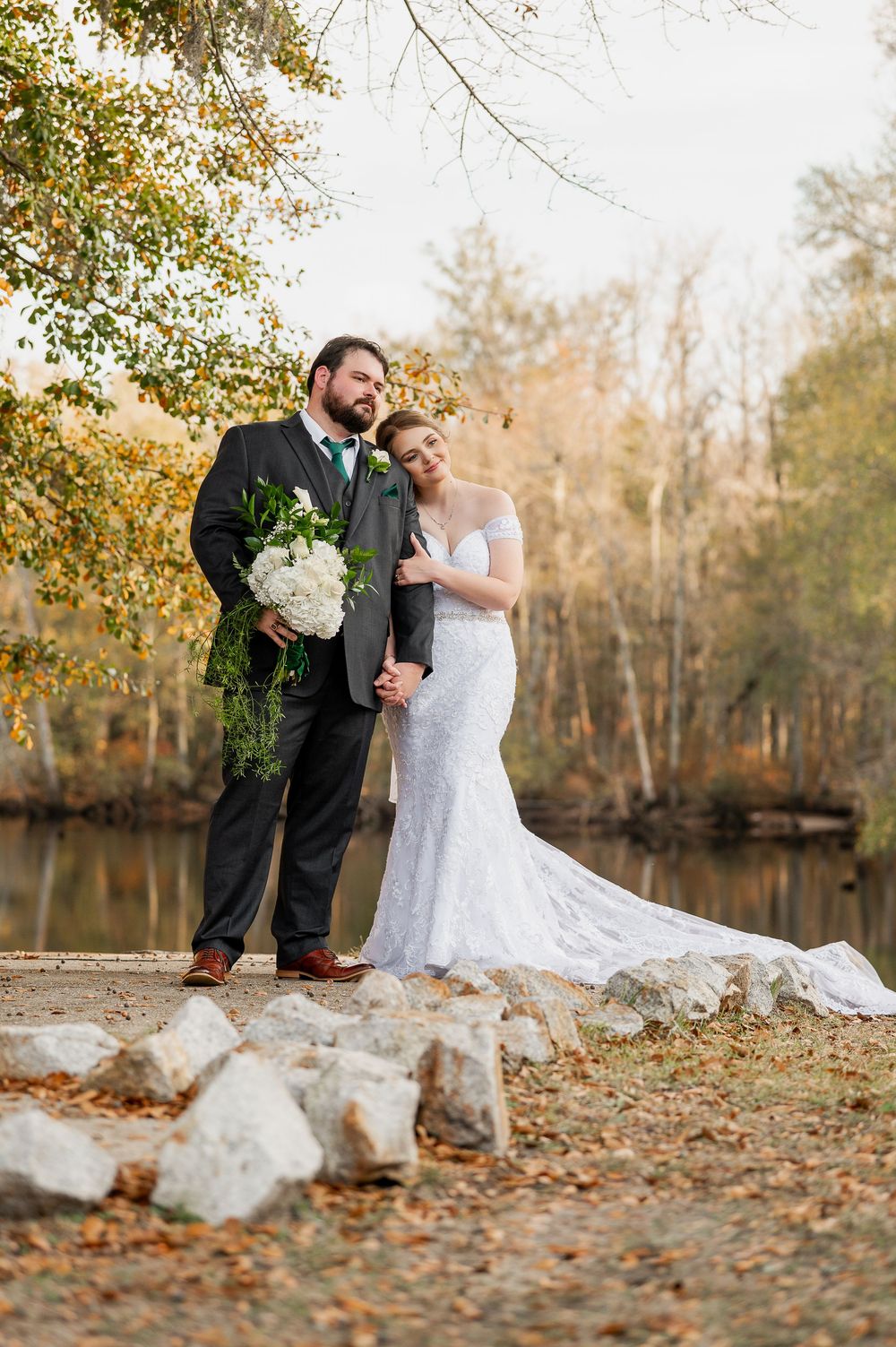 Newlyweds pose near large rocks on a leaf-covered path in an autumn forest setting.
