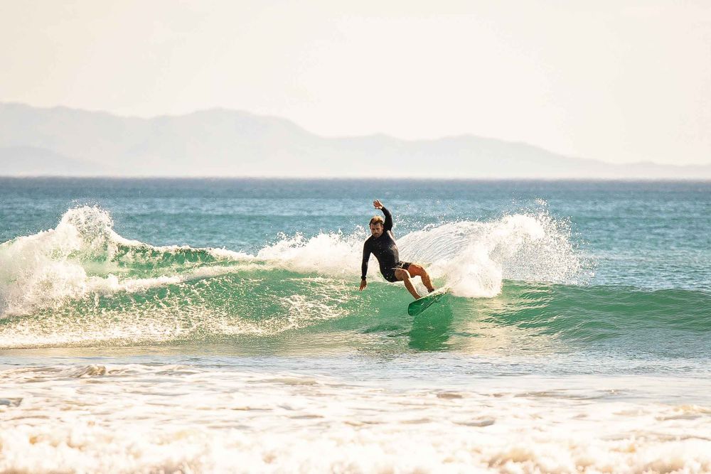 A surfer rides a green wave on a sunny day with mountains visible in the background along the coastline.