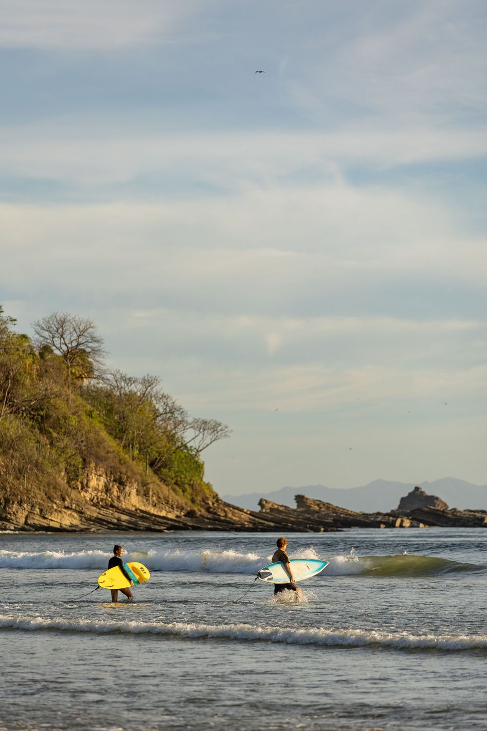 Surfers paddle out into the ocean waves near a rocky coastline during sunset with mountains in the distance.