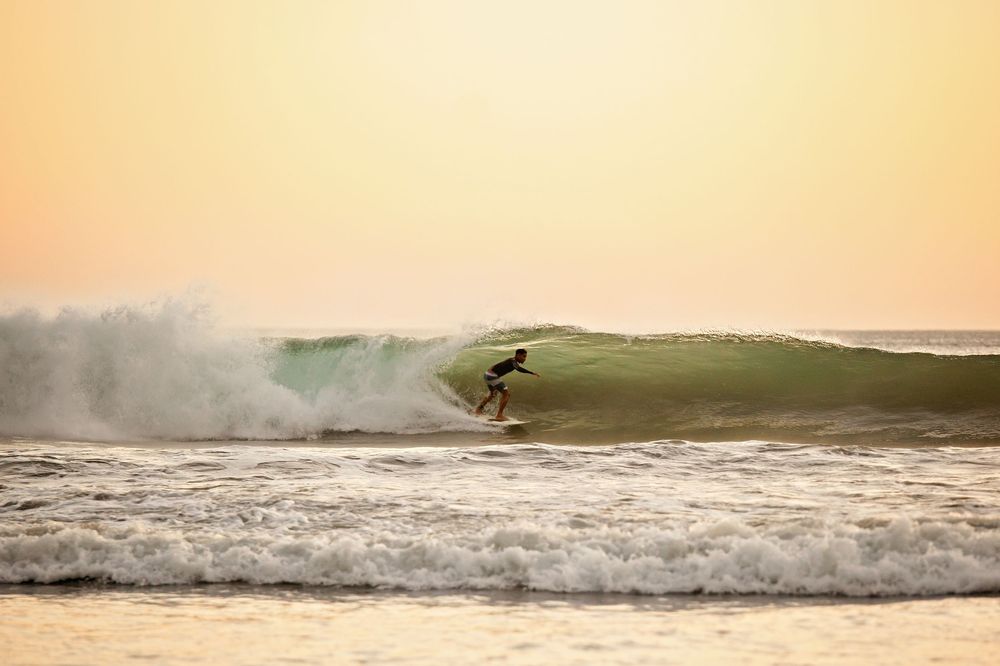 A surfer rides a wave at sunset with warm orange sky and breaking waves in the background.