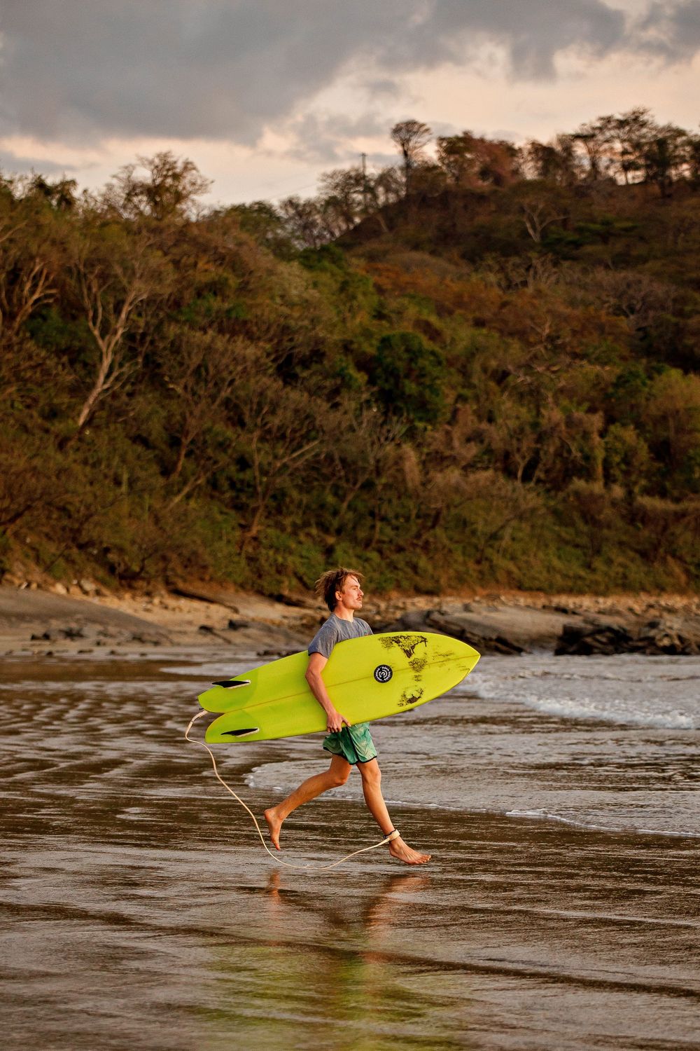 A surfer carries a bright yellow surfboard across a sandy beach at sunset with tropical trees in the background.