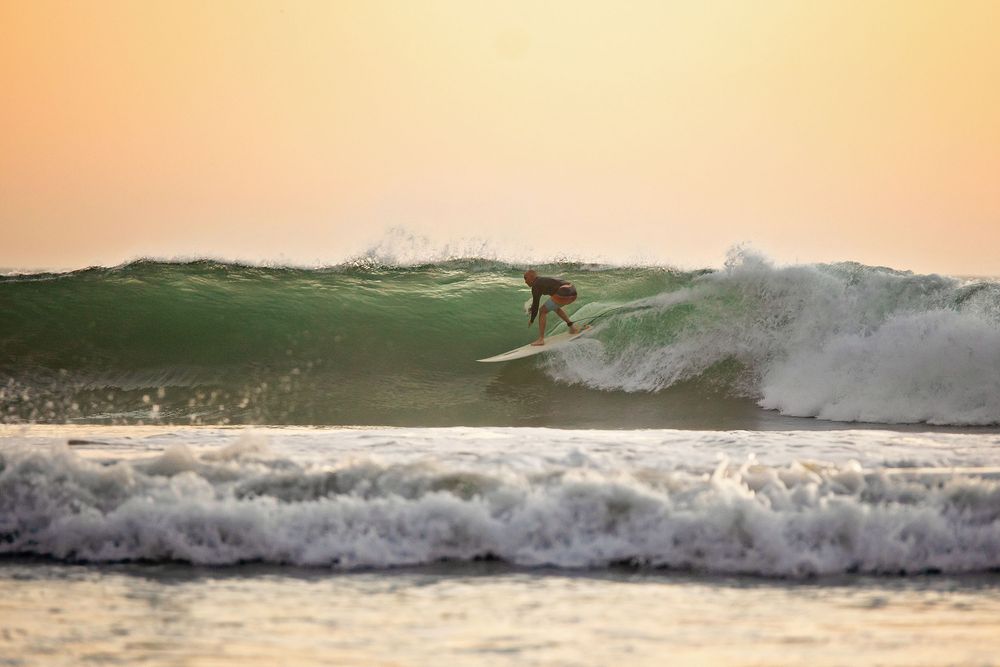 A sequence of ocean waves breaking at sunset with warm orange lighting and silhouettes visible in the surf.