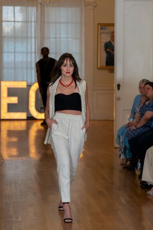 Fashion models showcase white and neutral summer outfits on wooden floor runway with glowing letters behind.