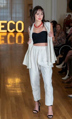 Model walks runway in white wide-leg pants and black crop top with ECO sign illuminated behind.