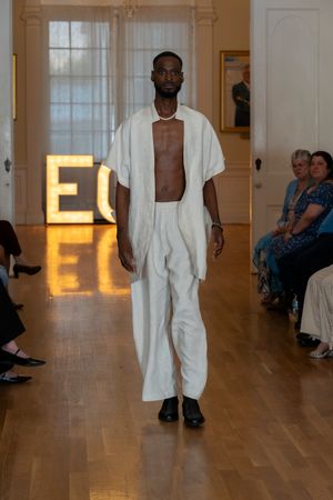 Model showcases minimalist white ensemble on wooden runway with illuminated backdrop.
