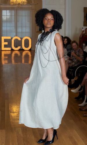 Model walks runway in flowy white dress with black necklace against illuminated ECU sign at fashion show.
