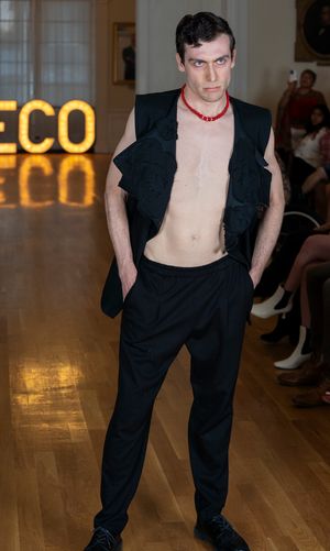 Model in black suit strikes dramatic pose on fashion runway with ECO sign illuminated behind.