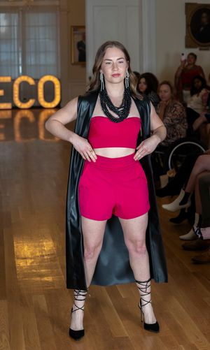 Model wearing red shorts and black jacket poses confidently on fashion runway.
