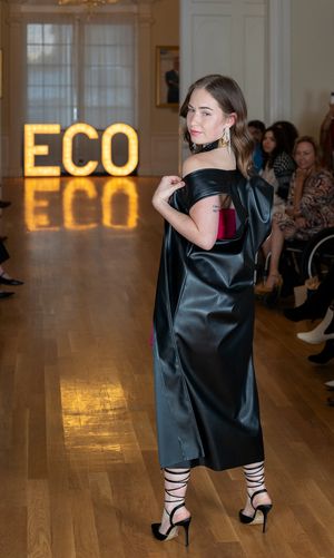 Fashion model walks runway in black satin dress with strappy heels against illuminated ECO sign at fashion show.