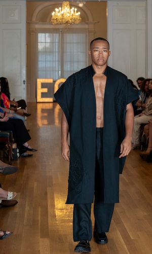 Model walks runway in oversized black ensemble during fashion show in elegant room with chandelier.