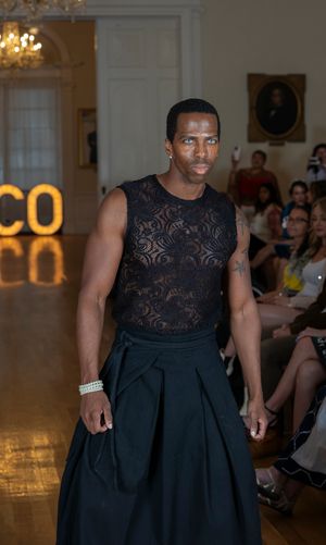 Model presents elegant black evening gown with lace bodice and pleated skirt on fashion runway.