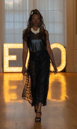 Model presents flowing black lace dress on runway with illuminated letters in background.