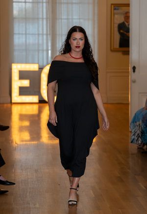 Model walks runway in flowing black dress at fashion show with illuminated letters in background.