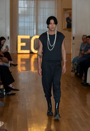 Fashion model walks runway in all-black ensemble with pearls during fashion show against illuminated ECO sign.