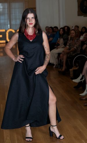 Model presents flowing black dress with pearl accessories on wooden runway with audience in background.