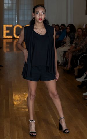 Model walks runway in black mini dress and strappy sandals at fashion show.