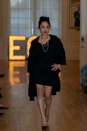Model struts down catwalk in sleek black ensemble with confident poses against illuminated backdrop.