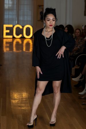 Model in little black dress and pearls poses on runway with illuminated ECO sign in background.