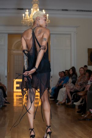 Model presents sheer black evening wear with strappy details on illuminated catwalk.
