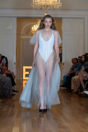 Model walks in ethereal sheer white tulle dress on runway beneath crystal chandelier.