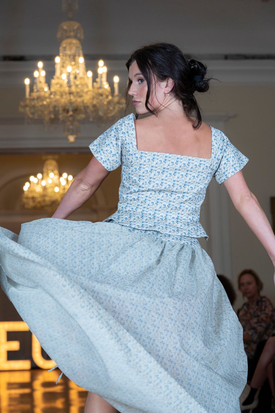 Model spins in light blue patterned dress on fashion runway with dramatic lighting.