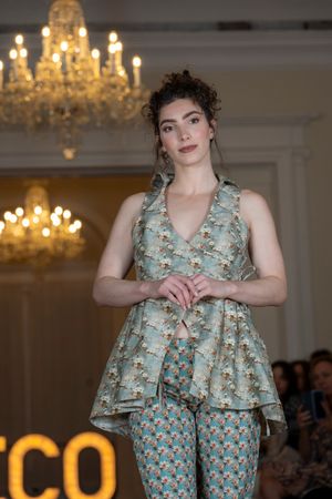 Runway model presents mint green floral coordinated outfit at fashion show with ornate chandeliers.