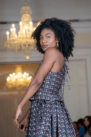 Fashion model poses in floral print dress during runway show with crystal chandeliers in background.