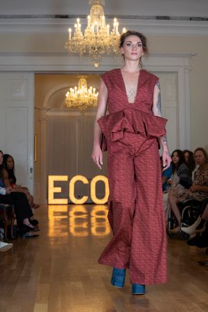 Model walks runway in burgundy textured pantsuit with peplum detail at eco fashion show under crystal chandeliers.