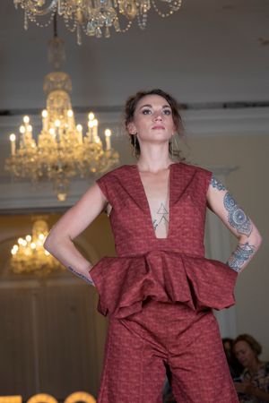 Model wearing a burgundy textured sleeveless dress poses under crystal chandeliers in a fashion show sequence.