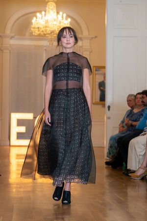 Model walks runway in sheer black cocktail dress with ECO signage visible.