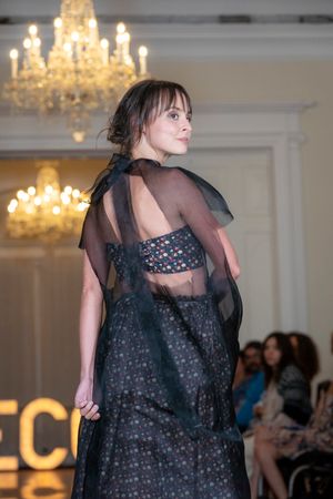 Model displays sheer black evening gown with cutout details on runway beneath crystal chandelier.