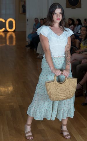 Model walks runway in floral mint dress with white top and straw handbag at fashion show.