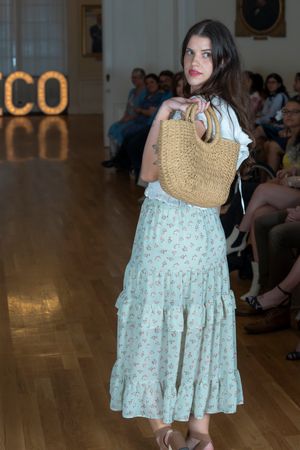 Model showcases floral print dress and straw bag on elegant runway with chandeliers.