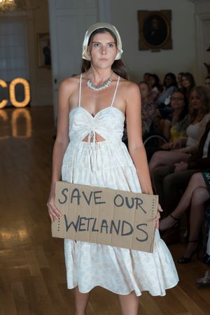 Model in white dress holds environmental protest sign about wetlands conservation during fashion show.