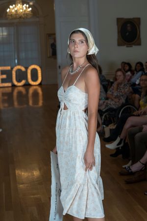 Fashion model walks runway in white summer dress with headscarf at fashion show with ECO signage and chandelier.
