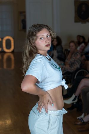 Fashion model in white cropped shirt poses dramatically on catwalk with lit signage behind.
