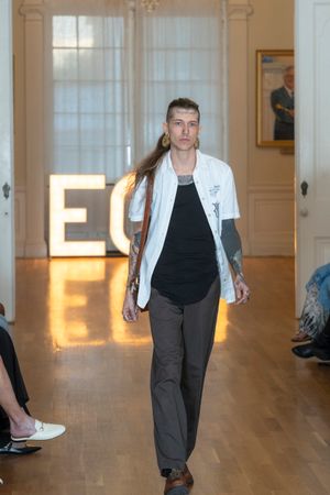 Model wearing white jacket and dark pants walks fashion runway with ECO lighting.