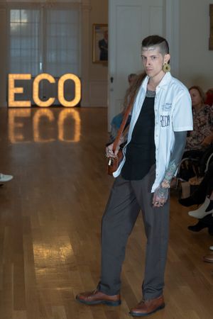 Model in white jacket and dark pants walks fashion runway with ECO sign glowing in background.