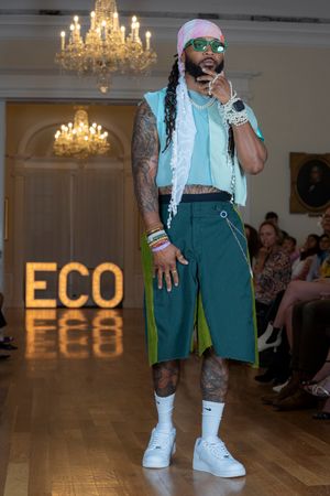 Fashion model wearing mint green outfit with shorts and bandana walks runway under crystal chandeliers at ECO fashion show.