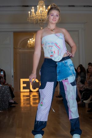 Model presents patchwork denim and fishnet fashion design on illuminated catwalk.
