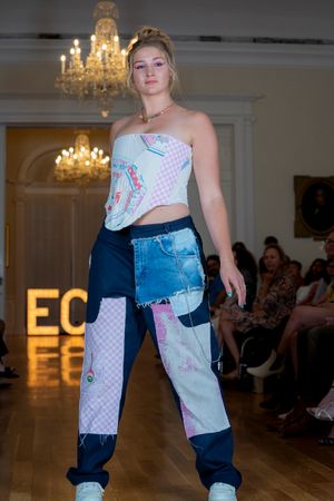 Model presents patchwork denim pants with mixed patterns and textures on indoor runway.