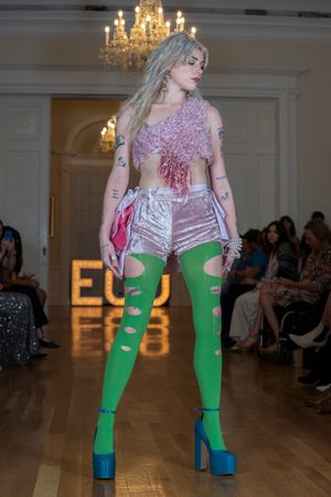 Fashion model walks runway in sparkly pink dress with bright green cutout tights and blue platform shoes under chandelier.