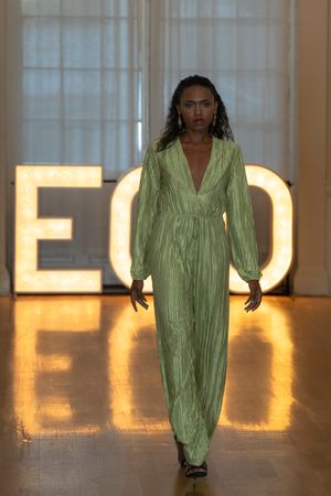 Sage green pleated jumpsuit with plunging neckline shown on illuminated runway at fashion event.