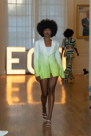 Model presents ombre green and white outfit on illuminated runway with audience in background.