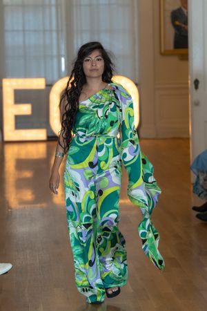 Model showcases flowing green and white abstract print maxi dress on runway with illuminated backdrop.