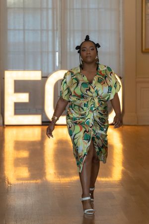 Model struts in vibrant green and yellow patterned dress on illuminated runway.