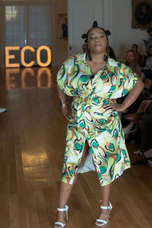 Fashion model showcases tropical print dress with white sandals on catwalk.