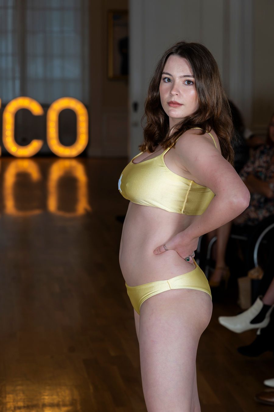 Model wearing yellow lingerie set poses on runway with illuminated 'CO' sign visible in background.