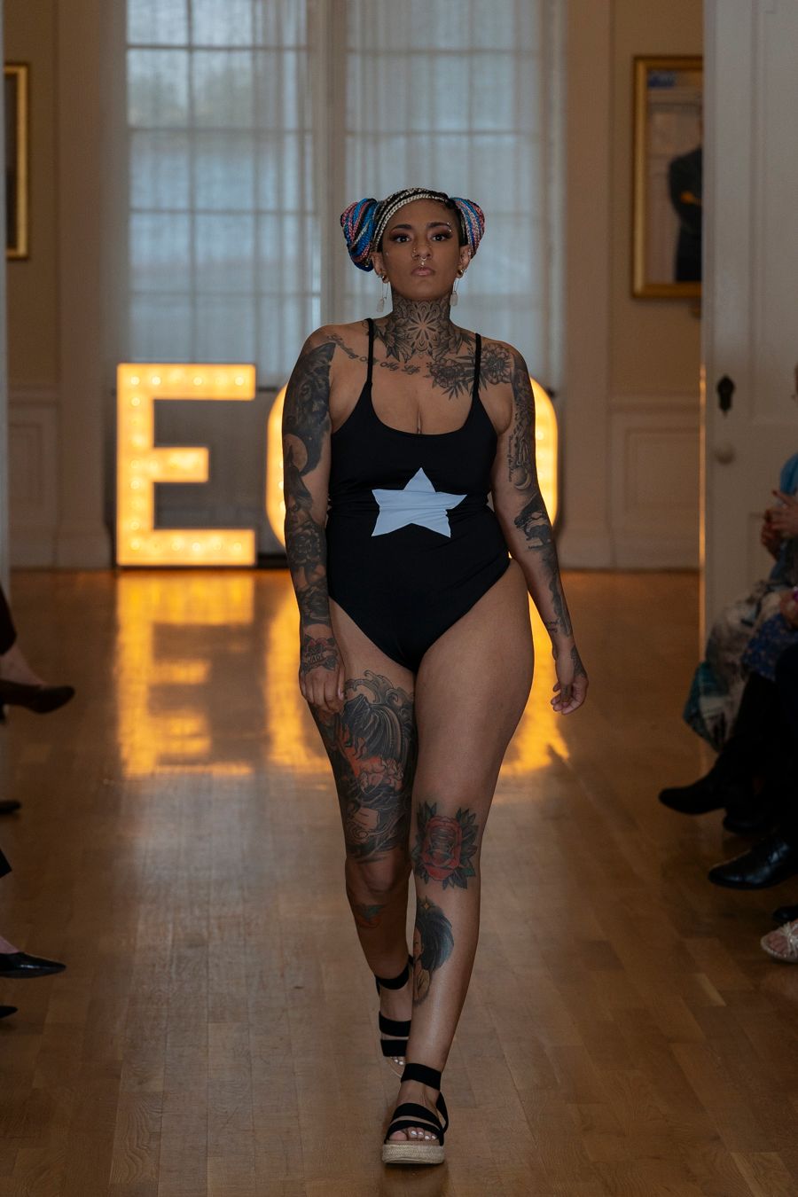 Model walks runway in black swimsuit with star design and sandals during fashion show in warmly lit hallway.